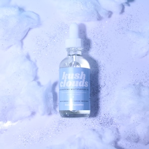 May include: A clear glass bottle with a dropper top and a white label that says "kush clouds detoxifying multipurpose oil" with the text "negative energies flee, touch me, vibrate higher" on the bottom of the label. The bottle is sitting on a white surface with blue and silver glitter.