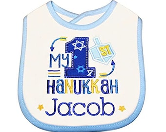 May include: A white baby bib with a blue trim. It features an embroidered design that says "My 1st Hanukkah Jacob" with a blue number one, a Star of David, and a dreidel.