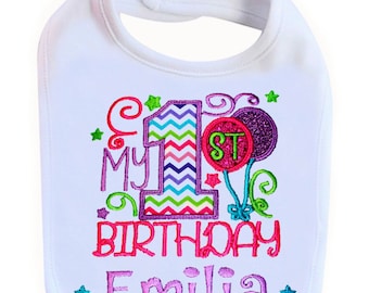 May include: White bib with colorful embroidered text that reads "My 1st Birthday Emilia". The number one is decorated with a chevron pattern and the text is surrounded by stars and swirls.