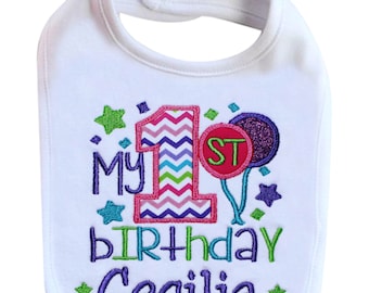 May include: White bib with colorful embroidered text that reads "My 1st birthday Cecilia" with a pink, purple, and blue chevron pattern on the number one, green stars, and a purple balloon with a red "ST" on it.