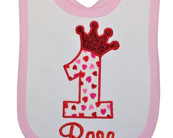 May include: White baby bib with a pink trim. The bib has a red number one with a pink and red heart pattern and a red glitter crown on top. The name "Rose" is embroidered below the number one.