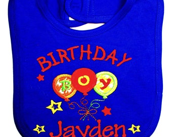 May include: Blue baby bib with the text "Birthday Boy" and the name "Jayden" in red. The bib has three red, yellow, and green balloons with the letters "BOY" on them.