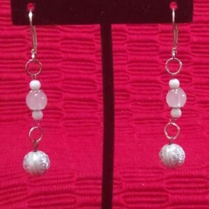 May include: Silver-tone dangle earrings with a series of white and pink beads and a textured silver-colored ball at the bottom. The earrings hang from a silver hook and are displayed against a red background.