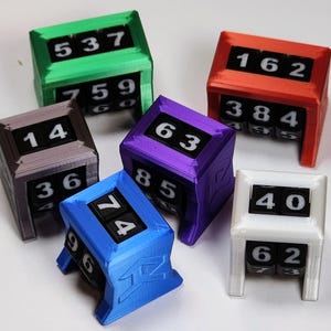 May include: A collection of six mechanical life counters in green, red, purple, blue, white, and grey. Each rectangular plastic frame houses rotating black number wheels with white digits, displaying various values like 537, 162, 14, 63, 74, and 40.