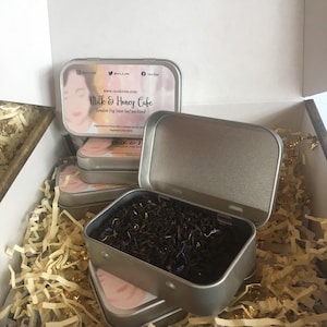 May include: A silver tin with a label that reads "Milk & Honey Cake London Fog loose leaf tea blend" and a picture of a woman. The tin is open and filled with loose leaf tea.