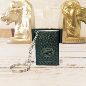 May include: A black book keychain with a silver key ring. The book cover features the title "Moby Dick or The Whale" by Herman Melville. The cover has a wave pattern and a whale illustration.