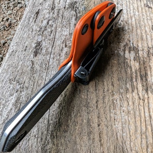 May include: A black and silver knife with an orange leather sheath. The sheath has three black snaps and a black clip.
