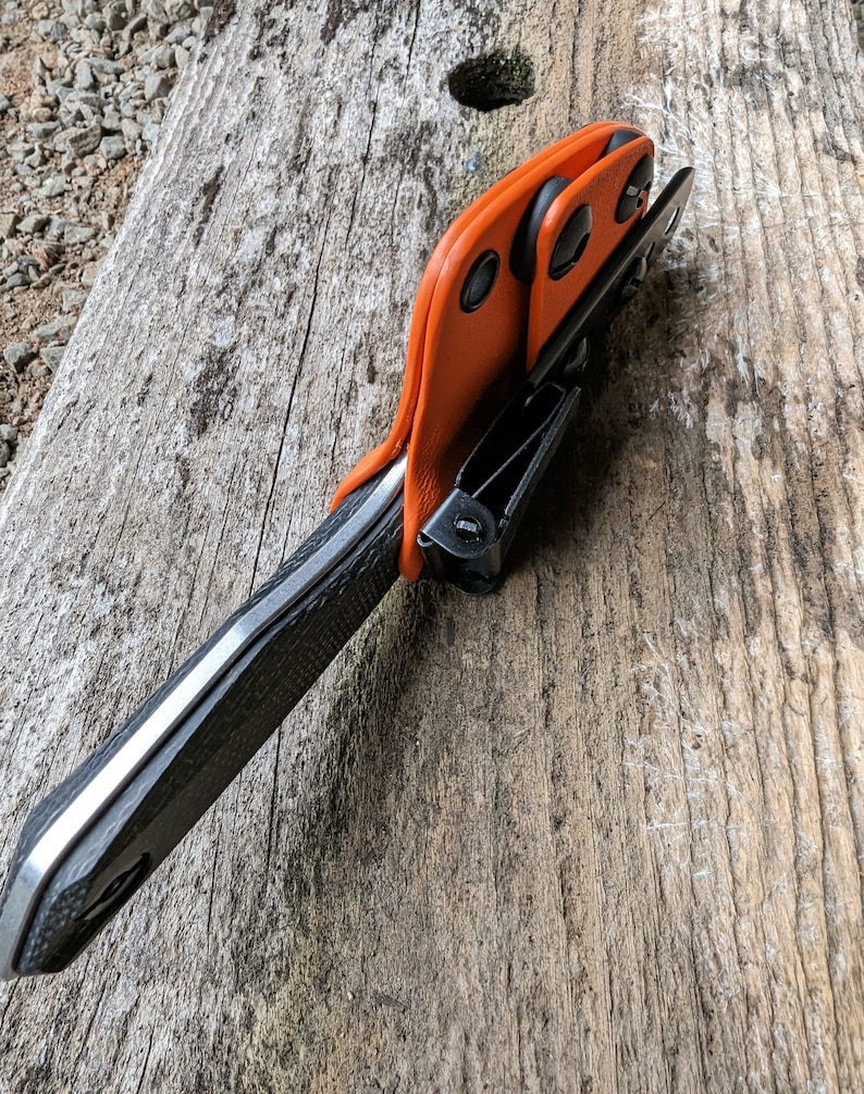 May include: A black and silver knife with an orange leather sheath. The sheath has three black snaps and a black clip.