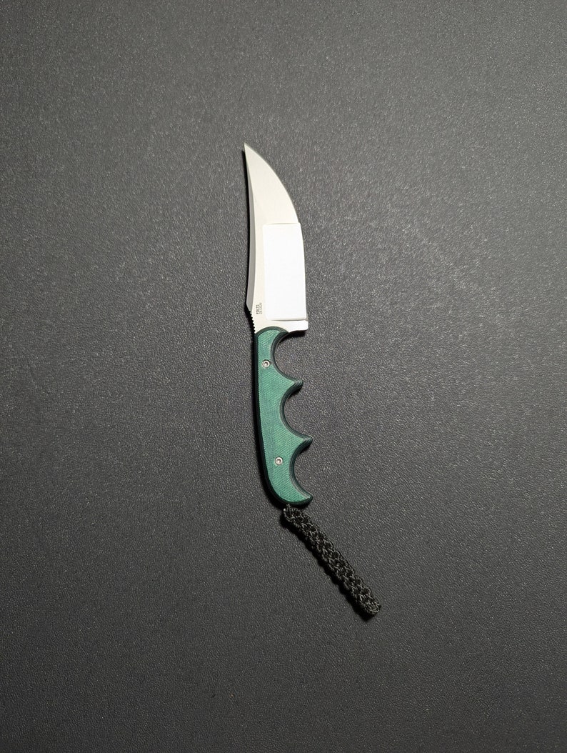 Puede incluir: Un cuchillo con mango verde, hoja plateada y un cord&oacute;n de lani&egrave;re negro. La hoja es curva y el mango tiene ranuras para los dedos.