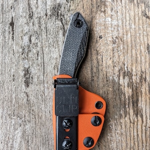 May include: A black knife with a black and orange leather sheath. The sheath has a black clip with the text "ULTI CLIP" on it.