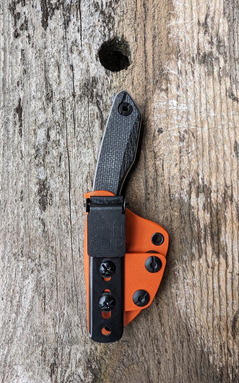 May include: A black knife with a black and orange leather sheath. The sheath has a black clip with the text "ULTI CLIP" on it.