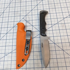 Puede incluir: Un cuchillo Gerber con mango negro y hoja plateada. El cuchillo está en una funda de cuero naranja brillante con un clip negro. El cuchillo y la funda están sobre una cuadrícula negra y blanca.