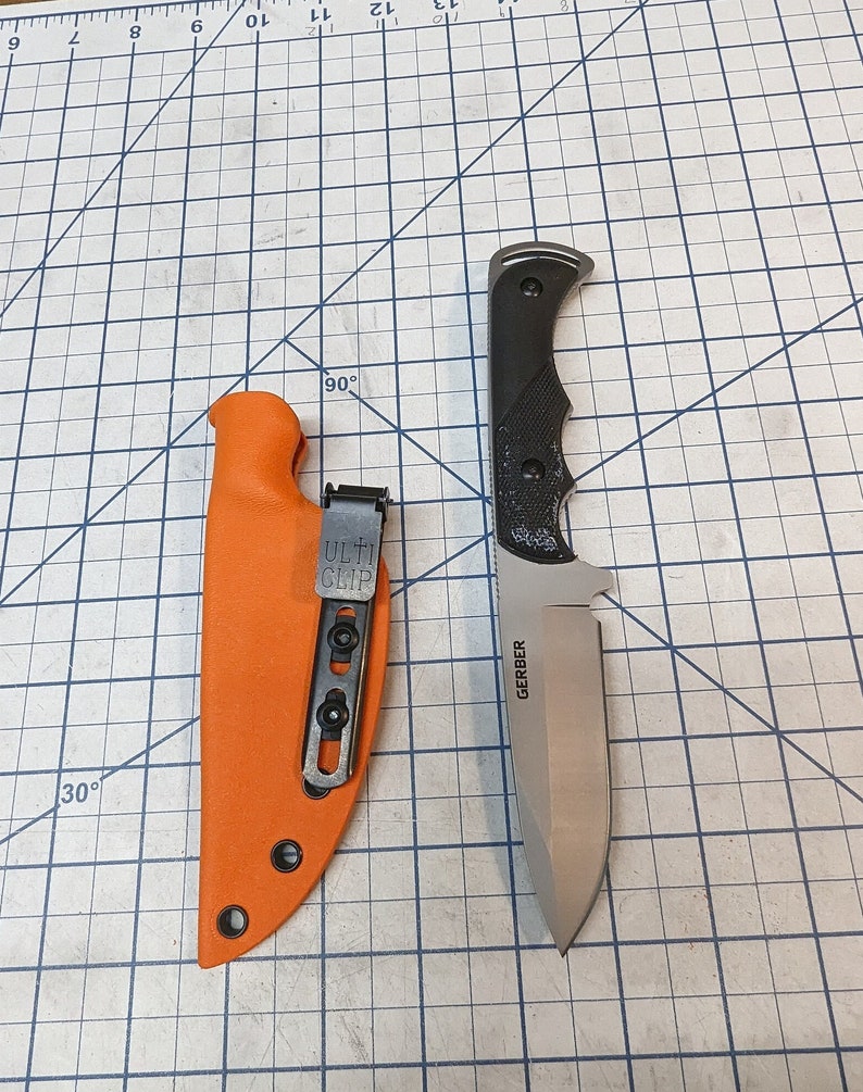 May include: A Gerber knife with a black handle and a silver blade. The knife is in a bright orange leather sheath with a black clip. The knife and sheath are on a black and white grid.
