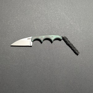 Puede incluir: Un peque&ntilde;o cuchillo de bolsillo negro y verde con un cord&oacute;n. El cuchillo tiene una hoja parcialmente dentada y un mango con ranura para los dedos. El nombre de la marca "CRKT" est&aacute; grabado en la hoja.