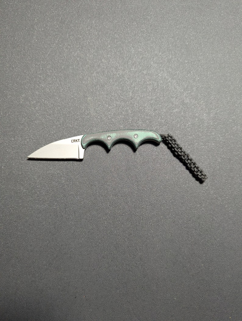 Puede incluir: Un peque&ntilde;o cuchillo de bolsillo negro y verde con un cord&oacute;n. El cuchillo tiene una hoja parcialmente dentada y un mango con ranura para los dedos. El nombre de la marca "CRKT" est&aacute; grabado en la hoja.