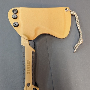 May include: A tan Gerber hatchet with a black and tan handle, secured in a tan sheath. The sheath is fastened with three black screws and a camouflage lanyard. The hatchet is designed for outdoor use.
