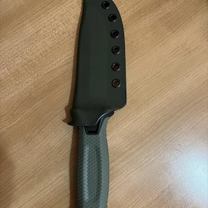 Puede incluir: Un cuchillo verde con mango negro y una funda verde con varios agujeros. El cuchillo tiene un mango texturizado y una hoja negra.