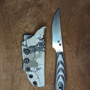 May include: A silver knife with a black and white patterned handle and a camo patterned sheath.