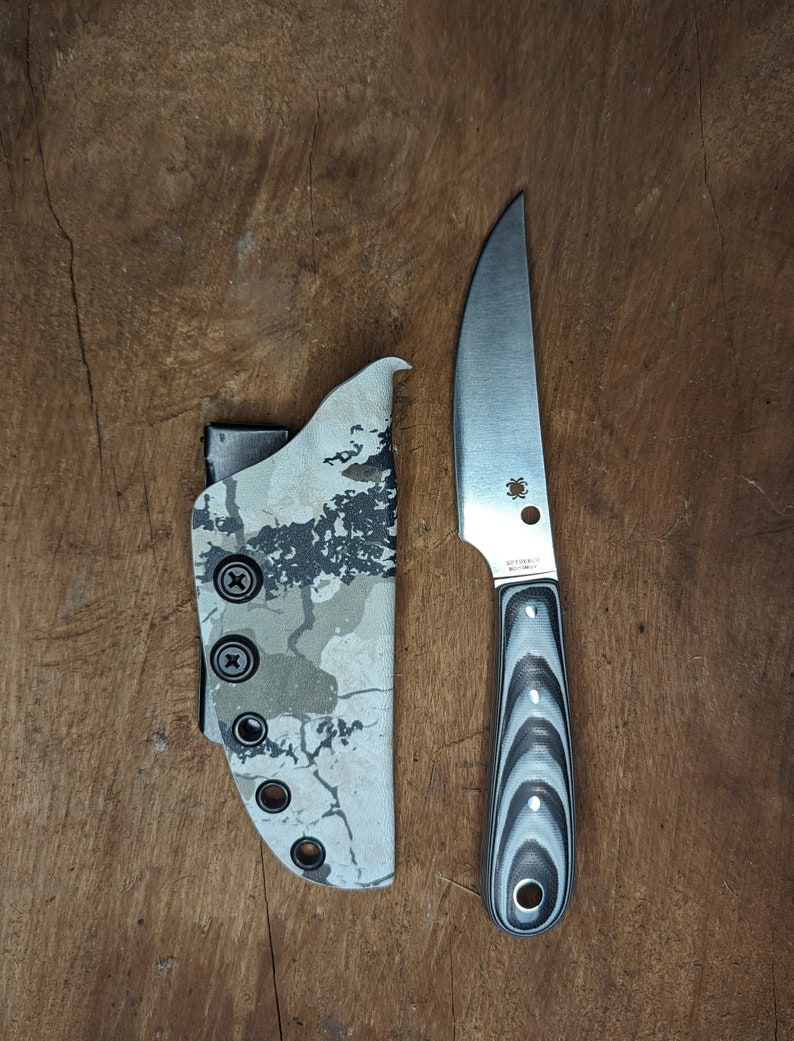 May include: A silver knife with a black and white patterned handle and a camo patterned sheath.