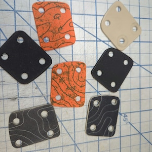 May include: A set of seven square-shaped, black and orange rubber pads with four holes each. The pads have a textured surface and are arranged on a gridded surface.