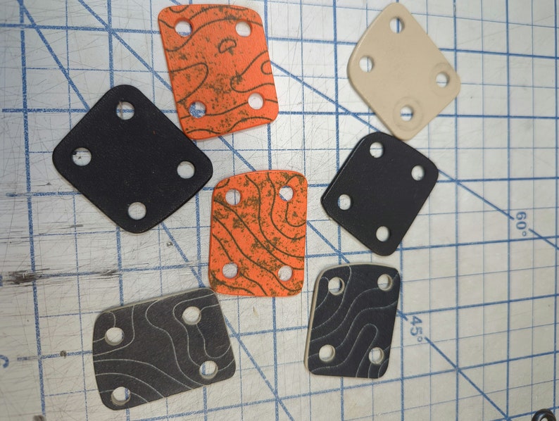 May include: A set of seven square-shaped, black and orange rubber pads with four holes each. The pads have a textured surface and are arranged on a gridded surface.