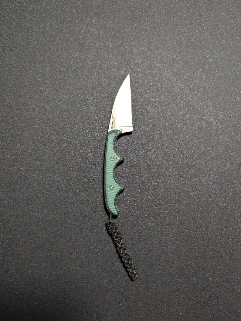 Puede incluir: Un cuchillo de bolsillo verde y negro con una hoja curva y un cord&oacute;n. El cuchillo tiene un mango con ranura para los dedos y el texto "MINIMALIST" est&aacute; impreso en la hoja.