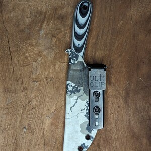 May include: A camo patterned knife sheath with a black and white patterned handle and a black Ulti Clip attached.