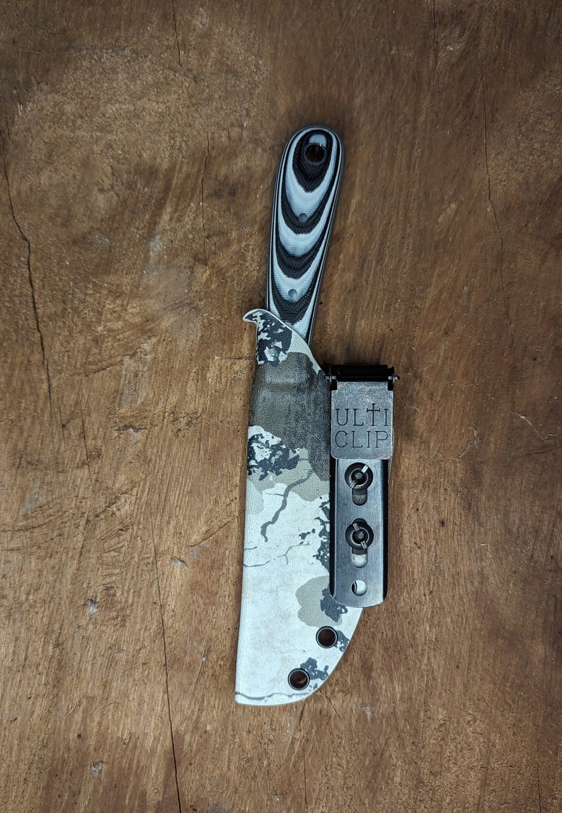 May include: A camo patterned knife sheath with a black and white patterned handle and a black Ulti Clip attached.