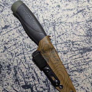 Puede incluir: Un cuchillo negro y verde oliva con el nombre de la marca "MORAKNIV" impreso en el mango. El cuchillo est&aacute; en una funda marr&oacute;n con un dise&ntilde;o de mapa topogr&aacute;fico y un clip de cintur&oacute;n negro. El cuchillo est&aacute; dise&ntilde;ado para uso en exteriores.