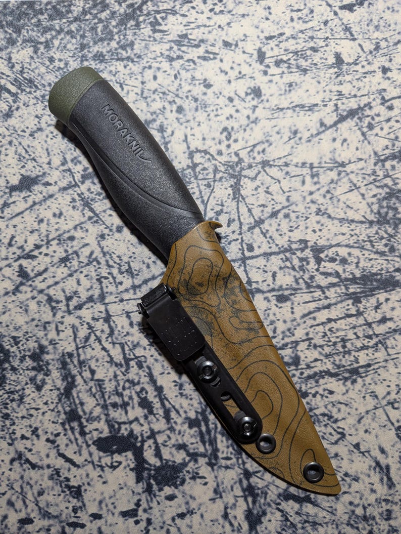 Puede incluir: Un cuchillo negro y verde oliva con el nombre de la marca "MORAKNIV" impreso en el mango. El cuchillo est&aacute; en una funda marr&oacute;n con un dise&ntilde;o de mapa topogr&aacute;fico y un clip de cintur&oacute;n negro. El cuchillo est&aacute; dise&ntilde;ado para uso en exteriores.