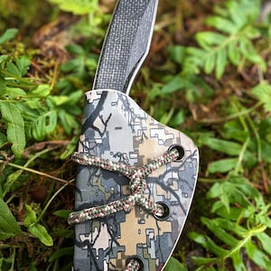 May include: A black knife with a carbon fibre handle is in a camouflage patterned sheath with a brown and green cord.