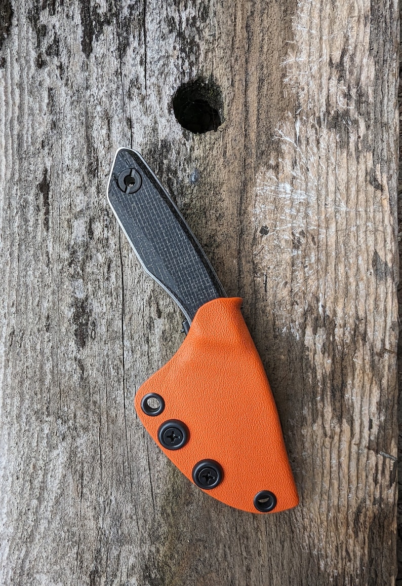 May include: A black and gray pocket knife with a carbon fiber handle and an orange leather sheath. The sheath has three black snaps.