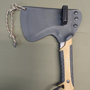 May include: A black and tan Gerber axe with a black sheath and a tan handle. The axe has a lanyard attached to the handle and a black clip on the sheath.