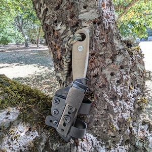 May include: A black leather knife sheath with a brown handle knife attached. The knife is secured to the sheath with a black clip. The sheath is attached to a tree trunk with a green and brown paracord.