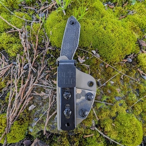 May include: A black knife with a carbon fiber handle is attached to a brown and green camouflage patterned belt clip. The clip has the text "Belt Clip" printed on it.