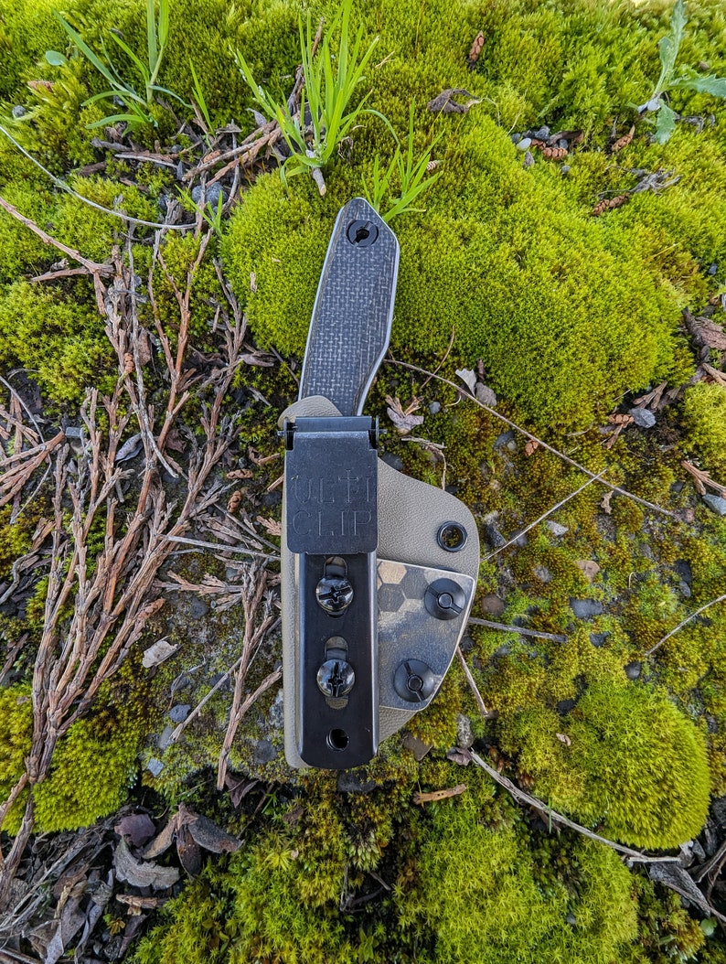 May include: A black knife with a carbon fiber handle is attached to a brown and green camouflage patterned belt clip. The clip has the text "Belt Clip" printed on it.