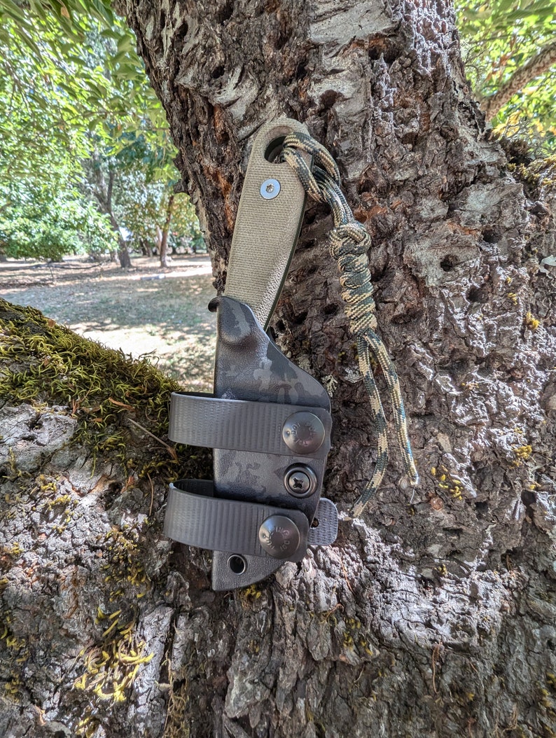 May include: A black and green camouflage knife sheath with a brown leather handle and a black nylon strap. The sheath is attached to a tree trunk with a green and brown rope.