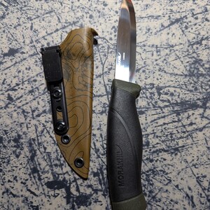 Puede incluir: Un cuchillo de acero inoxidable con mango negro y verde y funda marr&oacute;n. La hoja del cuchillo es brillante y afilada. La funda tiene un patr&oacute;n topogr&aacute;fico. El mango tiene la inscripci&oacute;n "MORAKNIV".