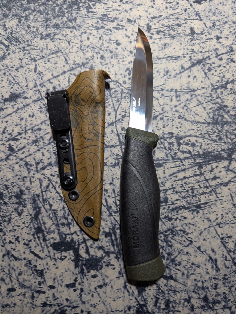 Puede incluir: Un cuchillo de acero inoxidable con mango negro y verde y funda marr&oacute;n. La hoja del cuchillo es brillante y afilada. La funda tiene un patr&oacute;n topogr&aacute;fico. El mango tiene la inscripci&oacute;n "MORAKNIV".