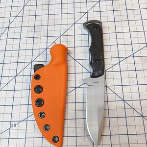 May include: A black handled knife with a silver blade and a black and orange sheath. The knife has a 4600723A serial number.