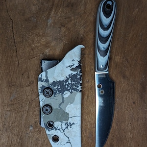 May include: A black and silver knife with a white and black patterned handle. The knife is in a camouflage patterned sheath.