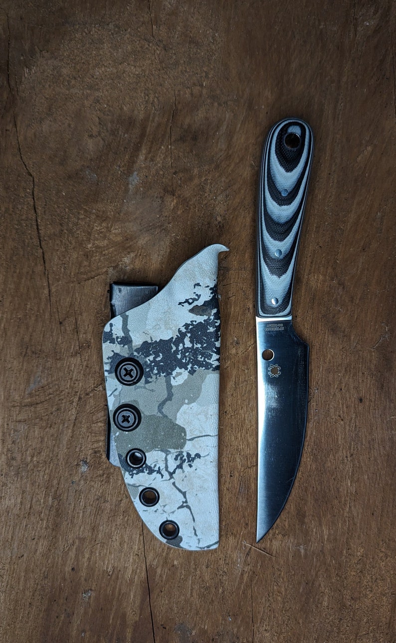 May include: A black and silver knife with a white and black patterned handle. The knife is in a camouflage patterned sheath.