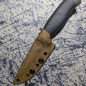 Puede incluir: Un cuchillo negro y verde con una funda con patr&oacute;n topogr&aacute;fico. El mango es negro con un extremo verde. La funda es de color canela con un dise&ntilde;o topogr&aacute;fico negro y cierres negros.