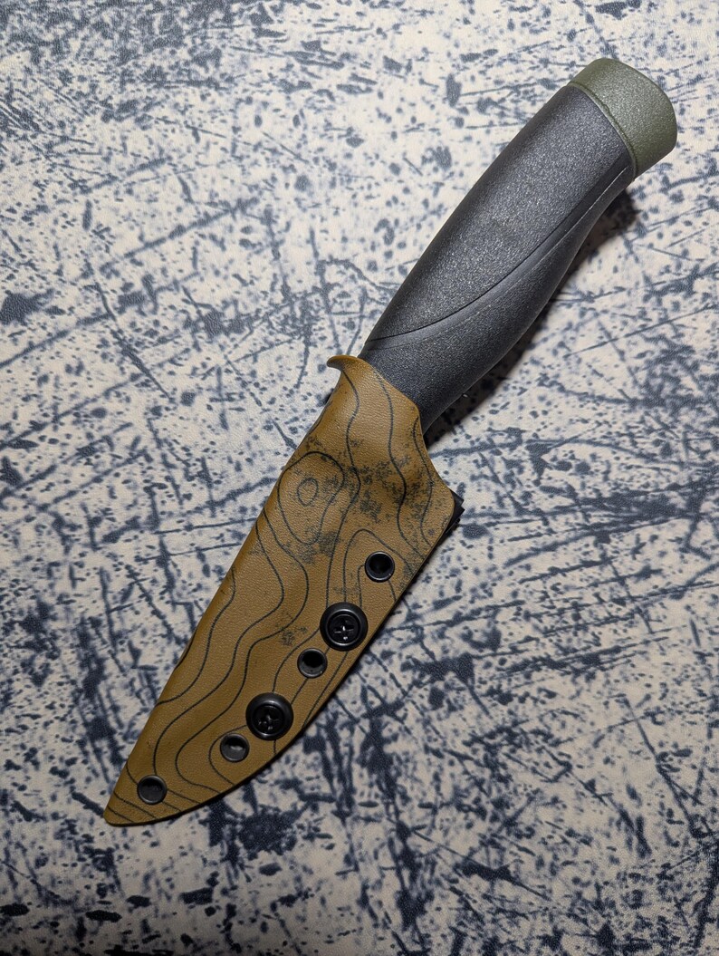 Puede incluir: Un cuchillo negro y verde con una funda con patr&oacute;n topogr&aacute;fico. El mango es negro con un extremo verde. La funda es de color canela con un dise&ntilde;o topogr&aacute;fico negro y cierres negros.