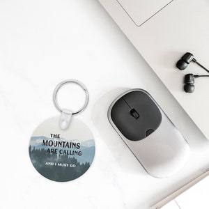 Może przedstawiać: Okrągły brelok do kluczy z niebieskim i białym motywem górskim i tekstem "The Mountains Are Calling And I Must Go".