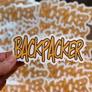 Op de afbeelding: Een stapel gele en witte "Backpacker" stickers. De stickers zitten in een hand, met de bovenste sticker die omhoog wordt gehouden.