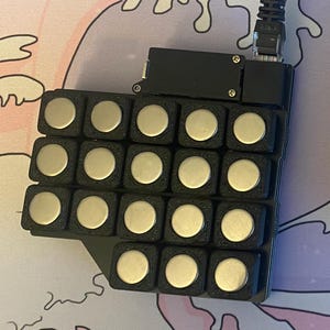 Puede incluir: Dos dispositivos electrónicos rectangulares negros conectados por un cable. Cada dispositivo tiene una cuadrícula de 25 botones redondos plateados.