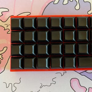 Può includere: Una tastiera compatta e rettangolare con tasti neri e una cornice rossa. La tastiera è posizionata su uno sfondo colorato con un design a onde rosa, bianco e viola.