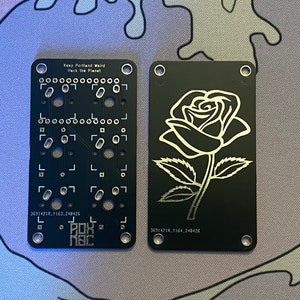 Puede incluir: Dos placas de circuito impresas negras con texto y diseños plateados. La placa de la izquierda tiene el texto "Keep Portland Weird Hack the Planet" y un diseño de placa de circuito impreso. La placa de la derecha tiene un diseño de rosa plateado. Ambas placas tienen cuatro agujeros en las esquinas.