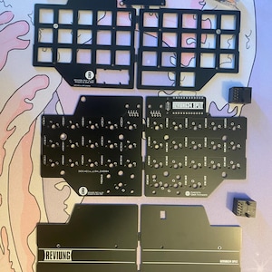 Könnte beinhalten: Schwarze Metallplatten für einen benutzerdefinierten Tastaturaufbau. Die Platten sind mit "REVUING 64 SPLIT" beschriftet und verfügen über ein Lochraster für Schalter und Tastenkappen. Die Platten sind für ein geteiltes Tastaturlayout ausgelegt.